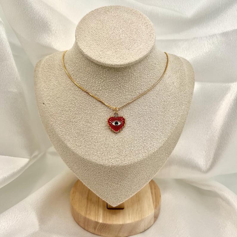 May include: A gold-colored necklace with a heart-shaped pendant. The pendant is red with a black eye design in the center. The necklace is displayed on a mannequin with a wooden base, set against a white fabric backdrop.