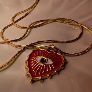 May include: Gold-colored necklace with a heart-shaped pendant. The pendant is red with a white eye design and gold accents. The chain is a thin, snake chain style.