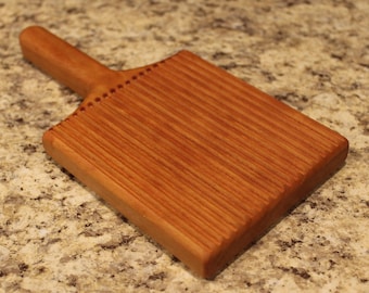 May include: A wooden butter paddle with a handle and a grooved surface. The paddle is made of light brown wood and has a reddish-brown hue.