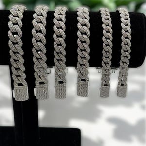 Peut inclure: Six bracelets de chaîne cubaine argentés et glacés sont présentés sur un présentoir en velours noir. Chaque bracelet présente un design de chaîne épais et imbriqué et un fermoir sécurisé. Les bracelets sont présentés sur un fond vert flou.