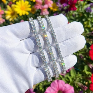 May include: Three silver tennis bracelets, each adorned with a row of sparkling, clear gemstones. The bracelets are displayed on a white glove, with a vibrant floral background of yellow, pink, and red flowers.
