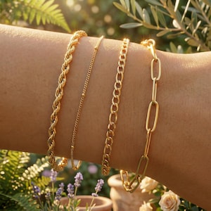 Può includere: Quattro braccialetti d'oro di vari design adornano un braccio. I braccialetti includono una catena a corda, una delicata catena di perline, una catena a maglia e una catena con maglie ovali allungate. I braccialetti sono esposti in un ambiente naturale all'aperto con vegetazione.