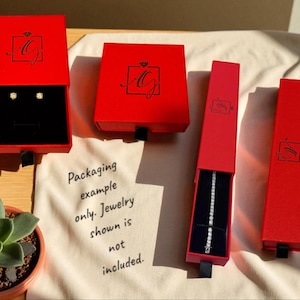 Puede incluir: Cajas rojas para joyas con cintas negras, algunas abiertas para revelar interiores de terciopelo negro. Una caja contiene pendientes, otra una pulsera. Las cajas est&aacute;n marcadas con un logotipo. Una peque&ntilde;a planta suculenta en primer plano.