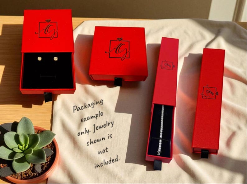 Puede incluir: Cajas rojas para joyas con cintas negras, algunas abiertas para revelar interiores de terciopelo negro. Una caja contiene pendientes, otra una pulsera. Las cajas est&aacute;n marcadas con un logotipo. Una peque&ntilde;a planta suculenta en primer plano.
