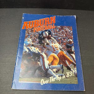 May include: A blue Auburn Football program from 1983 featuring a photograph of players in action on the field. The cover displays the text AUBURN FOOTBALL in orange letters at the top and the phrase Over The Top in '83! in white at the bottom.