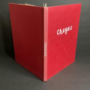 May include: A red book with the name "CHAGALL" in white script on the cover. The spine also features the name "CHAGALL" vertically. The book is open, revealing white pages.