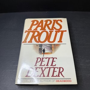 May include: A paperback book titled "Paris Trout" by Pete Dexter. The cover features the title in large orange letters, with an illustration of a man walking down a street. The author is also the author of "Deadwood".