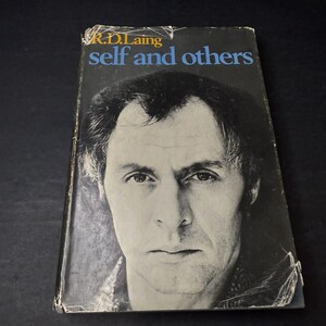 May include: A vintage book titled "self and others" by R.D. Laing. The book cover features a black and white portrait of a man with dark hair and a serious expression. The title is in blue and the author's name is in gold.