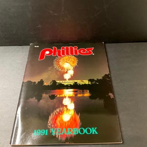 Op de afbeelding: Een Phillies-jaarboek uit 1991 met een nachtscène met vuurwerk boven water. Het woord "Phillies" staat in rode letters bovenaan en "1991 YEARBOOK" staat in groen onderaan.