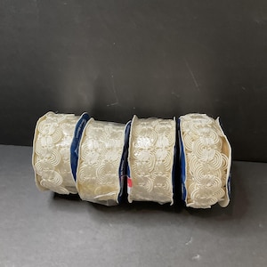 May include: Four rolls of cream-colored decorative ribbon with an intricate, repeating floral pattern. The ribbon is neatly rolled and held together with blue and white packaging. The background is a dark gray.