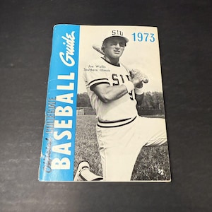 May include: A vintage 1973 Southern Illinois University (SIU) Collegiate Baseball Guide. The cover features a black and white photo of Joe Wallis, a baseball player, holding a bat. The guide is blue and white.
