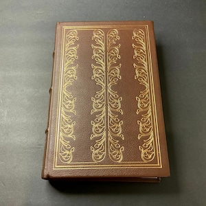 May include: A brown hardcover book with gold decorative filigree patterns. The book's cover features a rectangular design with vertical gold embellishments. The spine is visible, and the book is set against a dark background.