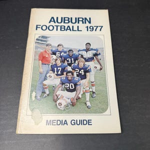 May include: A vintage Auburn Football 1977 Media Guide. The cover features a team photo of football players in blue and orange uniforms, with the coach in a red shirt. The guide is on a black surface.