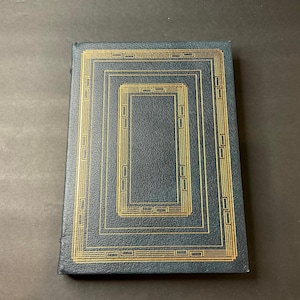 May include: A dark blue book with gold rectangular geometric designs. The cover features a series of nested rectangles with decorative detailing. The book's spine is visible, suggesting it is a bound volume. The overall aesthetic is classic and elegant.
