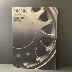 May include: A book titled "SAAB 9000 ENGINEERING FEATURES" with a grayscale image of a car wheel. The cover is a close-up of the wheel's design, with a focus on its metallic details and curved spokes. The background is a dark gray.