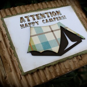 May include: A handmade card with a brown corrugated cardboard cover. The card has a white interior with the text "ATTENTION HAPPY CAMPERS!" and a brown and plaid tent cutout.