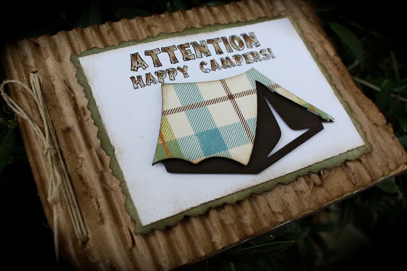 May include: A handmade card with a brown corrugated cardboard cover. The card has a white interior with the text "ATTENTION HAPPY CAMPERS!" and a brown and plaid tent cutout.