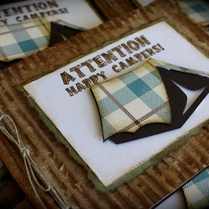 May include: A set of brown cardboard cards with a white paper insert. The insert has a green border and the text "ATTENTION HAPPY CAMPERS!" in brown letters. A brown and white plaid tent is on the insert.