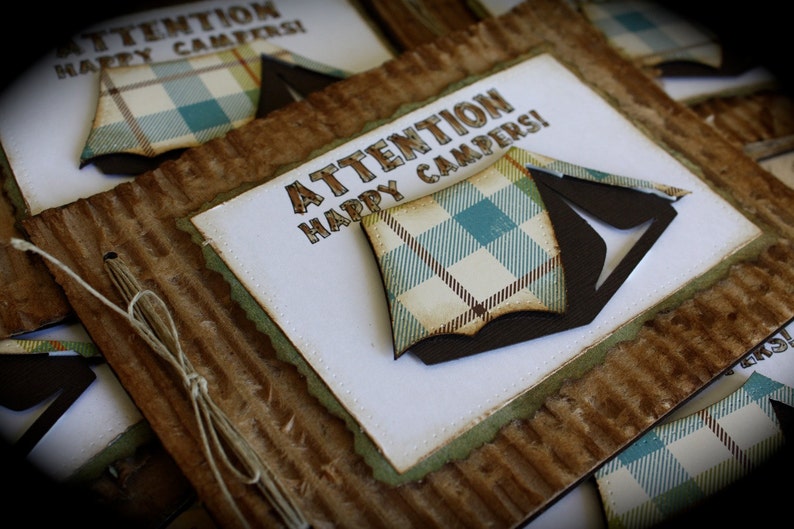 May include: A set of brown cardboard cards with a white paper insert. The insert has a green border and the text "ATTENTION HAPPY CAMPERS!" in brown letters. A brown and white plaid tent is on the insert.