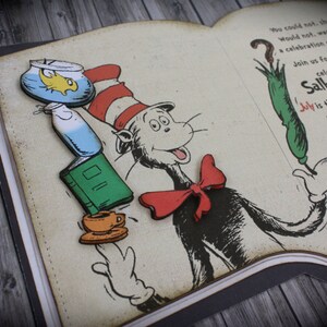 May include: A book-shaped invitation with a red bow, a green umbrella, a fishbowl, a book, and a cup of tea. The invitation is for Sally's 1st birthday party. The text on the invitation reads "Join us for a Seussical celebration for Sally's 1st Birthday. July is the month, the 7th is the day. The party starts at 11 so don't be late. Would you, come and celebrate with us? Please let us know. RSVP at..."