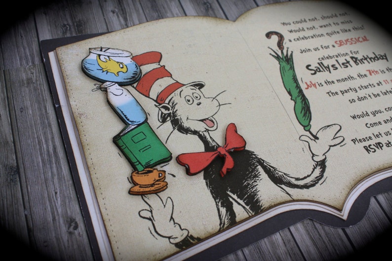May include: A book-shaped invitation with a red bow, a green umbrella, a fishbowl, a book, and a cup of tea. The invitation is for Sally's 1st birthday party. The text on the invitation reads "Join us for a Seussical celebration for Sally's 1st Birthday. July is the month, the 7th is the day. The party starts at 11 so don't be late. Would you, come and celebrate with us? Please let us know. RSVP at..."