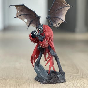 Puede incluir: Una figura de acción de Spawn que representa una criatura oscura y alada con rostro esquelético, dientes afilados y ojos verdes brillantes. La figura lleva una capa roja hecha jirones y guanteletes con púas sobre una base de lápida gris desgastada.