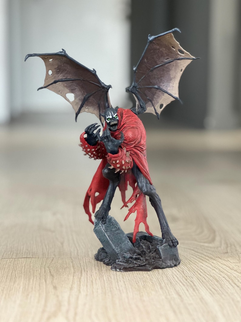 K&ouml;nnte beinhalten: Eine Spawn-Actionfigur, die eine dunkle, gefl&uuml;gelte Kreatur mit skelettartigem Gesicht, scharfen Z&auml;hnen und leuchtend gr&uuml;nen Augen darstellt. Die Figur tr&auml;gt einen zerfetzten roten Umhang und Stachelhandschuhe auf einem grauen, verwitterten Grabsteinsockel.