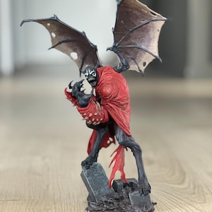 K&ouml;nnte beinhalten: Eine Spawn-Actionfigur, die eine dunkelgraue Kreatur mit rotem Umhang, stacheligen Handschuhen und gro&szlig;en, zerfetzten Fl&uuml;geln zeigt. Die Figur steht auf einem Sockel, der wie ein Friedhof mit einem Grabstein mit der Aufschrift RIP gestaltet ist.