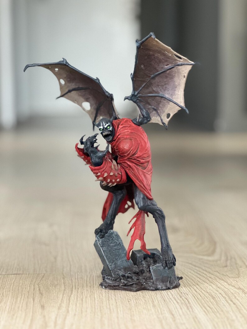 K&ouml;nnte beinhalten: Eine Spawn-Actionfigur, die eine dunkelgraue Kreatur mit rotem Umhang, stacheligen Handschuhen und gro&szlig;en, zerfetzten Fl&uuml;geln zeigt. Die Figur steht auf einem Sockel, der wie ein Friedhof mit einem Grabstein mit der Aufschrift RIP gestaltet ist.