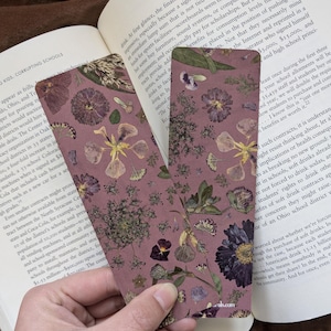 Puede incluir: Dos marcapáginas rectangulares con un fondo malva están adornados con flores prensadas de varios colores. Los marcapáginas se sostienen abiertos sobre un libro abierto. Los marcapáginas tienen un diseño floral.