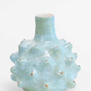 May include: A light blue ceramic vase with a textured surface and small, gold-colored accents. The vase has a bulbous body and a narrow neck, suitable for displaying flowers or as a decorative piece. The vase is approximately 8 inches tall.