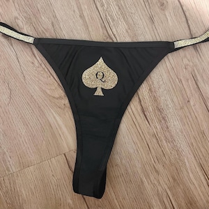 May include: Black thong underwear featuring a gold glitter spade symbol with the letter "Q". The straps are decorated with gold glitter. The item is shown on a wooden surface.