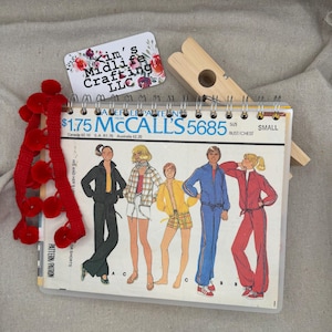 May include: A McCall's 5685 sewing pattern booklet in size small, featuring illustrations of five people in casual sportswear. A red pom-pom trim is draped over the left side, and a wooden clothespin rests on top, next to a Kim's Midlife Crafting LLC card.
