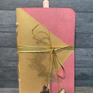 May include: A pink and gold journal with a gold ribbon tied around it. The journal has a gold leaf design on the front cover and two small keys hanging from the ribbon.