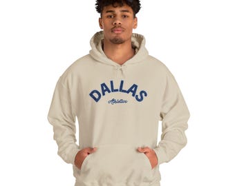 May include: A sand-coloured hooded sweatshirt featuring the text DALLAS in a large, arched, dark blue distressed font, with the word Athletics written in a smaller, cursive dark blue script directly underneath the main text on the chest.