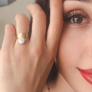 Peut inclure: Une bague en or avec un charme de perle blanche qui pend. La bague est portée à l'index d'une main.