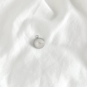 Könnte beinhalten: Ein kleiner, runder Silberanhänger mit dem Buchstaben "A" in der Mitte. Der Anhänger hat oben eine Schlaufe für eine Kette und liegt auf einem weißen Stoffhintergrund.