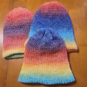 May include: Three knitted beanies in a rainbow gradient of colors, from red to yellow to blue. The beanies are handmade and appear soft, with a textured knit pattern. The hats are displayed on a wooden surface.