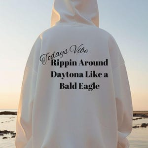 May include: A white hooded sweatshirt with the text "Todays Vibe Rippin Around Daytona Like a Bald Eagle" in black. A silhouette of a flying eagle is in the background.