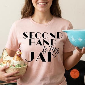 Puede incluir: Camiseta rosa claro con el texto negro "SECOND HAND IS MY JAM". La imagen también presenta un cuenco turquesa con diseños de estrellas blancas y un recipiente de cerámica con forma de gallina.