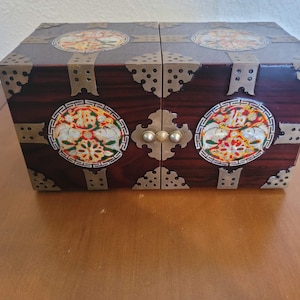 May include: A wooden box with a dark reddish-brown finish, adorned with decorative metal accents and circular, colorful designs. The box has a hinged, double-door design with a central clasp.