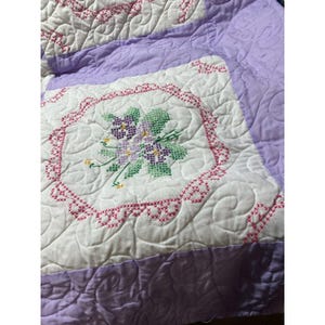 May include: A quilted bedspread with a floral cross-stitch design. The quilt features white squares with embroidered purple and green flowers, framed by a pink border. The edges and borders are a soft lavender color.
