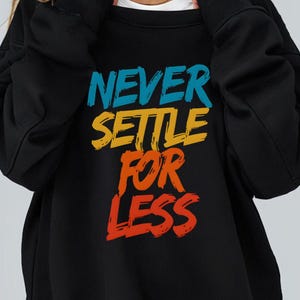 Può includere: Felpa nera con la scritta "NEVER SETTLE FOR LESS" stampata in lettere colorate e stilizzate.