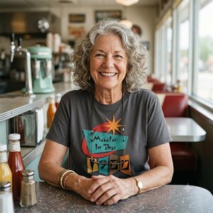 May include: A charcoal grey short-sleeved t-shirt featuring a graphic design with a teal trapezoid, a red oval, and an orange starburst. The text reads Made In The 1960s in black lettering. The shirt is worn by a person with curly grey hair.