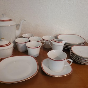 May include: A white porcelain tea set with red trim and gold accents. The set includes a teapot, sugar bowl, creamer, cups, saucers, and plates. The pieces are arranged on a wooden surface.
