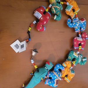 May include: A colorful beaded garland featuring stuffed animal figures in red, green, blue, and orange. The garland includes a bell and a tag with text. The figures are connected by a string of beads in various colors.