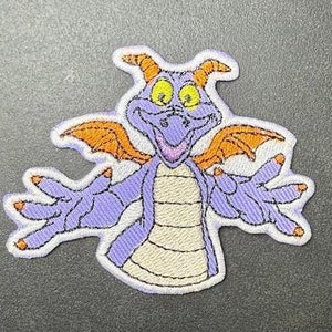 Könnte beinhalten: Gestickter Aufnäher mit einem lila Drachen mit orangefarbenen Flügeln und gelben Augen.
