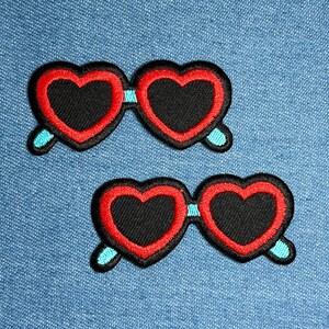 May include: Two embroidered patches of heart-shaped sunglasses. The lenses are black with a red outline, and the frames are red. The arms of the sunglasses are light blue. The patches are on a blue denim background.