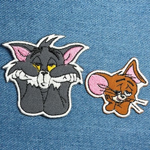 May include: Embroidered patches featuring Tom and Jerry cartoon characters. Tom is a gray cat with pink ears and yellow eyes, looking sad. Jerry, a brown mouse with pink ears, also appears dejected. Both patches have white borders and are set against a blue denim background.