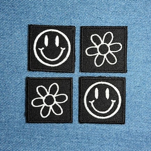 Könnte beinhalten: Vier quadratische schwarze Aufnäher mit weißen Stickereien. Zwei Aufnäher zeigen ein Smiley-Gesicht, die anderen beiden ein Blumenmuster. Die Aufnäher sind auf einem blauen Jeans-Hintergrund angeordnet.